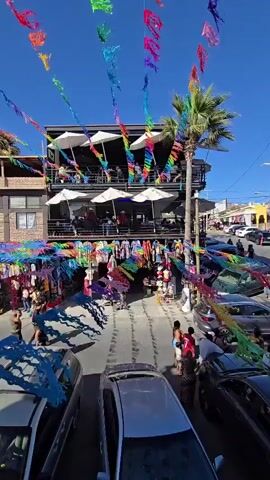 The video shows an overhead shot of a street market. The street is lined with stalls selling colorful textiles and clothing. A multi-story building with balconies and umbrellas is visible behind the stalls. There are people walking and browsing the market, and cars are parked on the sides of the street. Colorful banners are strung across the street, and a palm tree stands tall. The sky is clear and blue.