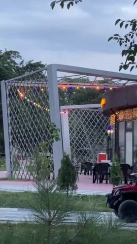 A young boy is riding a red toy car while a teenage boy pushes him from behind. A woman walks past in the background wearing a light-colored hijab and clothing. The scene takes place in an outdoor area with a modern, open-sided structure adorned with string lights. There are tables and chairs set up under the structure, suggesting an outdoor dining or seating area. Trees and greenery surround the area, and the sky appears overcast, indicating dusk or an evening setting.