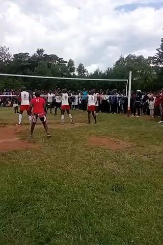 The video shows a volleyball game taking place on a grass field. Two teams are competing, with players wearing different color uniforms: one team in red and white, and the other in white and blue. A volleyball net is stretched across the middle of the field, separating the two teams. The background features a crowd of spectators watching the game, trees, and a cloudy sky. The camera angle is slightly shaky, providing a view from the sidelines. During the video, players are seen hitting and passing the ball, with one player diving to the ground to make a play.