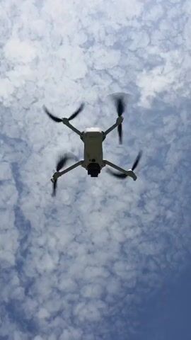 The video shows a drone ascending into a sky filled with puffy clouds. The drone is light-colored with four propellers that are visibly spinning. The camera angle is from directly below, looking up at the drone as it moves higher into the sky. The clouds provide a textured backdrop, and the blue of the sky peeks through in places. The drone becomes smaller as it gains altitude.