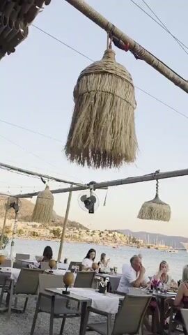This media file shows an outdoor restaurant with a view of the sea and a fleet of boats. The setting appears to be a coastal town, likely in Bodrum, Turkey, given the context provided. Several people are seated at tables, enjoying a meal or drinks. The atmosphere seems relaxed and social, with diners engaged in conversation.

The time of day is late afternoon or early evening, as indicated by the soft, warm light and the clear sky with a hint of twilight. The weather appears to be pleasant, with a gentle breeze suggested by the movement of the straw lampshades hanging from a bamboo structure above the tables. These lampshades are a distinctive decorative element, adding a rustic charm to the establishment.

In the background, a large cruise ship and numerous sailboats are visible on the water, suggesting a popular tourist destination. The coastline is dotted with buildings, and a historic structure, possibly a castle or fortress, can be seen in the distance, further hinting at the location's rich history and scenic beauty. A ceiling fan is mounted on the bamboo structure, likely for comfort during warmer periods.

The people in the video are diverse, and their attire suggests a casual yet elegant setting. Some are dressed in light summer clothing. The overall mood is one of leisure and enjoyment, characteristic of a vacation or a relaxed evening out. There is no visible text in the media file.