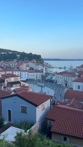 The video shows an aerial view of a European coastal town with red-tiled roofs and white buildings. The town is situated on a hill overlooking a harbor filled with boats and yachts. The sea stretches out to the horizon under a clear blue sky with a few wispy clouds. The scene appears to be a scenic overlook, possibly captured from a high vantage point.