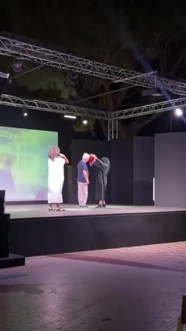 A stage is set outdoors at night. A screen displays a colorful, dreamlike image of clouds and arches. Three people stand on the stage. On the left, a person in a white robe with long, curly hair faces the screen. In the center, a person wearing a blue shirt and light-colored pants has a red and green wig. To the right, another person in a dark robe stands behind the person in the blue shirt, placing a hand on their shoulder and appearing to adjust their wig. The stage is lit with purple and white lights, and a metal truss structure with lights and a disco ball is visible above. Trees are in the background.
