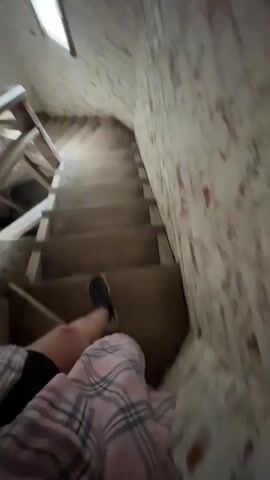 A first-person perspective video shows a person descending a narrow, winding wooden staircase. The walls are rough, textured, and appear to be made of exposed brick or plaster with a distressed finish. The staircase has wooden steps and handrails, some of which appear worn or unfinished. Light sources are visible at the top and possibly along the side of the stairs, casting a dim illumination. The person's legs, clad in plaid pajama pants and dark shoes, are visible as they navigate the descent. The overall environment suggests an attic, a basement, or an older, unfinished structure.