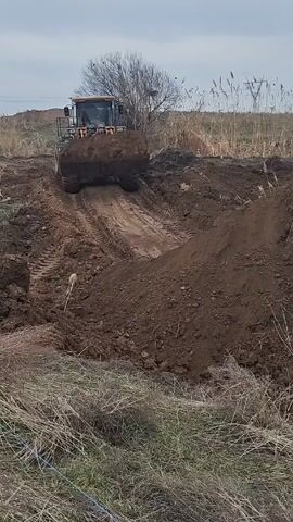 The scene shows a front-end loader moving a large scoop of dirt in a rural setting. The loader is positioned on a dirt track it has created, with piles of dirt mounded on either side. The ground is covered with dry grass and dirt. In the background, there's a tree with bare branches, reeds, and power lines. The sky is overcast, suggesting a cloudy day. The loader is orange and black and is angled slightly uphill, moving towards the viewer.