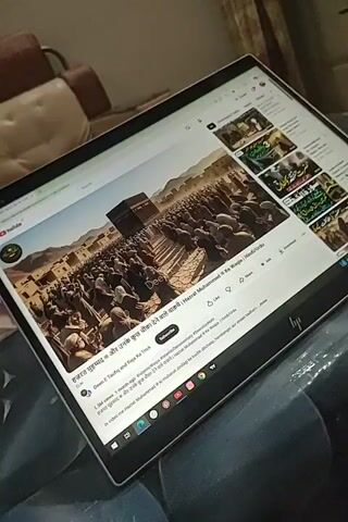 A laptop screen displays a YouTube video. The video thumbnail shows a large crowd of people surrounding the Kaaba in Mecca. The video title, visible on the screen, is in Hindi and appears to be about Prophet Muhammad. There are also other video thumbnails visible on the right side of the screen, some of which also seem to be related to Islamic content. The laptop is placed on a surface that appears to be a piece of furniture, possibly a couch or a bed, covered with a dark fabric.