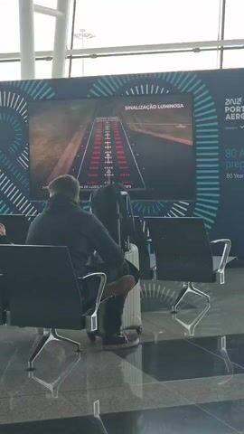 A static medium shot shows a man seated in an airport waiting area, viewing a large screen that displays an image of an airport runway illuminated with red and white lights. The man is seen from the back, wearing a dark jacket and dark pants, with his legs crossed and his head facing the screen. A carry-on suitcase and a backpack are positioned beside him. The airport's polished floor reflects the scene, including the chairs and the support structures of the building's architecture. The background includes a display with a blue and white patterned design, advertising ANA Aeroportos Portugal, with text in Portuguese about runway lighting.