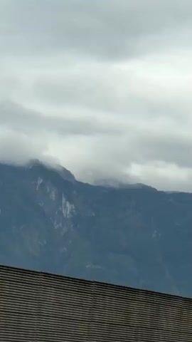 The video shows a view from a moving vehicle, likely on a highway. In the foreground, there is a wooden barrier. Beyond the barrier, there are trees and a mountain range covered in clouds. The sky is overcast. Several vehicles are visible on the road, including a white truck with "SteirerFleisch" written on its side. There is also an orange structure overhead with signs on it. The camera appears to be pointed upwards and slightly to the right.