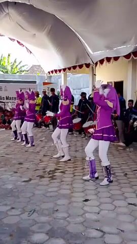 The video shows a group of girls in purple and gold marching band uniforms performing outdoors. They are wearing matching purple jackets with gold trim, white pants, and white boots with purple accents. Most of them are wearing purple head coverings. They appear to be dancing or marching in formation. Behind them, several people are playing drums. The performance is taking place under a large white tent with red trim. In the background, there is a building and some trees. The ground is paved with interlocking stones. There is an audience seated on the left side of the frame.