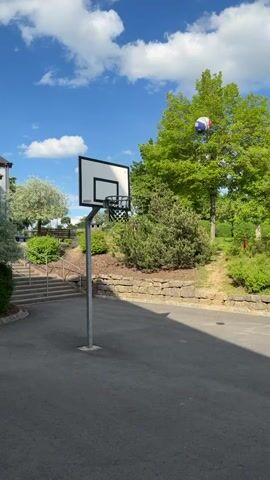 The video shows a man in a white shirt and black shorts playing basketball on an outdoor court. There is a basketball hoop in the foreground, surrounded by greenery and a bright blue sky with some clouds. The man picks up a ball and walks towards the hoop, dribbling it as he goes. The camera remains stationary, capturing the man's movements against the backdrop of the sunny day and lush landscape. The person is speaking in German, but it is not immediately apparent what is being said.
