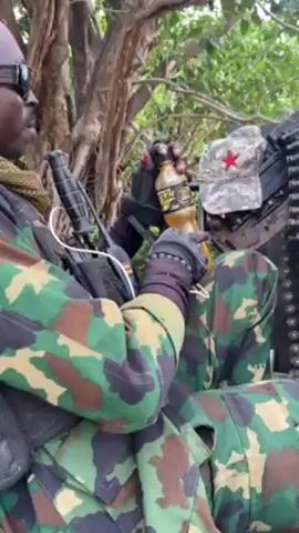 A person in military camouflage clothing is shown in an outdoor setting. The individual is wearing sunglasses and has what appears to be a headset attached. They are seated next to a large piece of military equipment, possibly a mounted weapon system, which has a cap with a red star placed on it. The person is holding a bottle of liquid and appears to be drinking from it. The background shows trees and greenery, suggesting the scene is in a wooded or rural area.