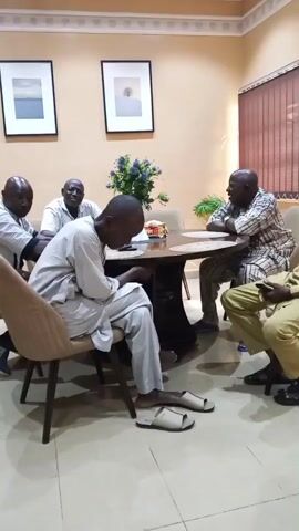 A group of six men are gathered around a dark wood table in a room with neutral-colored walls. Five of them are seated, while one stands. The seated men are dressed in a mix of casual and traditional attire, with one wearing a light gray outfit and sandals. He is looking down at an object in his hands. The other seated men are looking in different directions, seemingly engaged in conversation or observation. The standing man is wearing a tan shirt and is positioned near the table. The room is decorated with framed artwork on the walls and a plant arrangement on the table. Natural light filters in through a window with closed blinds.
