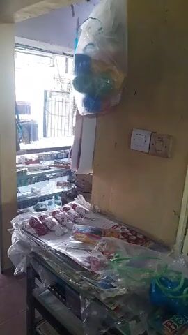 The video opens on a display shelf filled with various items, including what appear to be party favors or small toys in clear plastic bags. The items are brightly colored. Next, the camera pans to a large, beige copier machine. The room appears to be a shop or office. The next shot shows a woman wearing a red hijab and a patterned shirt sitting at a desk, using a computer. She is typing on the keyboard, and a printer is visible next to the computer. The shelves in the background are filled with various items, possibly stock for the shop. The lighting in the room is somewhat dim, with fluorescent lights visible on the ceiling. The overall impression is of a small business or service establishment.