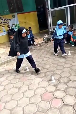 This video shows a group of children playing a game outdoors on a paved area. The children are wearing school uniforms, consisting of dark pants and light blue shirts with a darker blue stripe. Some of the girls are also wearing headscarves, in either light blue or black. The children are arranged in a line, facing towards the viewer. In front of the children, there are several white paper cups placed on the ground. One girl in the foreground, wearing a black headscarf, is shown in motion, appearing to be jumping or stepping on the cups. Other children are watching, and two adults, possibly teachers, are also present and observing the game. One adult, wearing a white garment and a red headscarf, is holding a microphone and seems to be addressing the children. The environment appears to be a schoolyard, with a building and some greenery visible in the background. The ground is paved with hexagonal tiles. The game seems to involve maneuvering around the cups on the ground.
