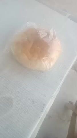 A close-up, slightly hazy overhead shot focuses on a single, round bread roll wrapped in clear plastic. The roll is golden brown on top and appears soft. It rests on a white, textured surface, possibly a tablecloth or placemat, which is laid out on a light-colored, possibly concrete or stone, ground. The background is out of focus, with a hint of a light-colored wall and some wooden elements, possibly furniture, visible on the right side. The lighting is soft and diffused.