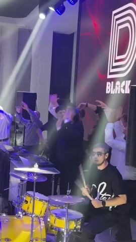 The video shows a live music performance in an indoor venue. A drummer is seated at a yellow drum set in the foreground, playing with drumsticks. A DJ setup with turntables and a laptop is positioned behind and to the left of the drummer. Several people are standing and dancing behind the DJ setup, with their arms raised. A large screen in the background displays a logo that appears to be "D BLACK STAR" in white and red lettering. Stage lights are casting beams of light onto the performers and the audience. The overall atmosphere appears to be energetic and celebratory.