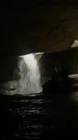 The video shows a cave-like environment with rocky formations and water.  In the first frame, a waterfall is visible pouring from an overhang into a pool of water.  In subsequent frames, the camera pans to reveal people standing in the cave.  The cave walls appear to be made of layered rock and are dark, with some areas illuminated by light.  There are at least three people visible in the cave, dressed casually. One person appears to be pointing at something. A triangular warning sign is visible on a rock ledge in one frame. The ground is covered in gravel and rock debris.