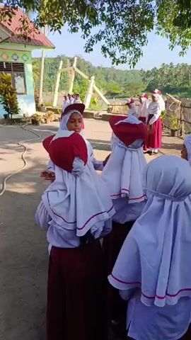 A group of schoolgirls in white and red uniforms are standing outdoors, likely rehearsing a performance. The girls are wearing white headscarves, with some having red accents on their headgear. They appear to be singing or reciting something, with one girl seemingly leading the group. The setting appears to be near a river or body of water, with lush greenery and trees visible in the background. A small building is also noticeable in the background, suggesting a school or public facility. The scene is captured during daylight, with clear and sunny weather.