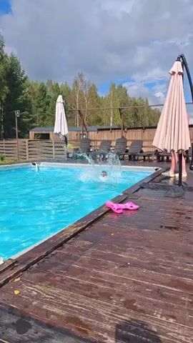A young person with blond hair splashes into a bright blue swimming pool from the left side of the frame. The person is mostly submerged, with their head and shoulders visible. Their arms are extended upwards, creating a splash. Pink flip-flops are visible on the wooden deck in the foreground, to the right of the pool. A wooden deck with a wet surface runs along the bottom of the frame. In the background, there are lounge chairs, umbrellas, a fence, and trees. The sky is cloudy. A person in red shorts is standing on the right side of the frame, partially visible.