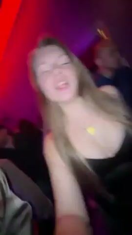 A young woman is filming a selfie video in a dimly lit nightclub or bar. She's wearing a black top and a gold necklace. She is holding a clear glass with a lime wedge in it, and sticks her tongue out at the camera. Behind her, a bald man in a blue shirt appears. The lighting is a mix of purple and red. The camera then turns and shows a crowd of people dancing. The video quality is somewhat shaky.