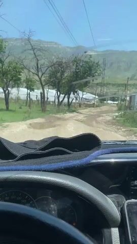 The video is shot from the interior of a vehicle, looking forward through the windshield. The dashboard and steering wheel are visible in the foreground, with a black cloth draped over the dashboard. Outside the vehicle, a dirt road stretches ahead, with puddles of water on the ground. The road is flanked by green grass and trees, some of which appear to be bare. In the background, there are several large, white, tent-like structures, possibly greenhouses. Further in the distance, rolling hills and mountains are visible under a bright blue sky with scattered clouds. Power lines stretch across the sky. The scene appears to be rural and somewhat undeveloped.