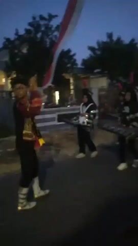 In a low-light video, a marching band performs on a street at dusk. The band members wear black uniforms with white vests and gold trim, and many are wearing hats with red plumes. Some members are playing drums, while others appear to be playing a glockenspiel. A large Indonesian flag, red and white, is visible in the background, attached to a pole. The street is paved, and there are houses and trees lining the sides. The overall atmosphere is one of a parade or performance. The video captures the band members marching in formation and playing their instruments. The lighting suggests it is either early morning or late evening.