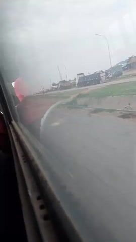 The video shows a view from inside a vehicle, looking out a window. Outside, there is a road with several large trucks parked or driving on it. The sky is cloudy. A person wearing a red shirt is visible in the foreground, partially obscured by the window. The overall impression is that of a journey or a stop along a roadside with heavy vehicles.