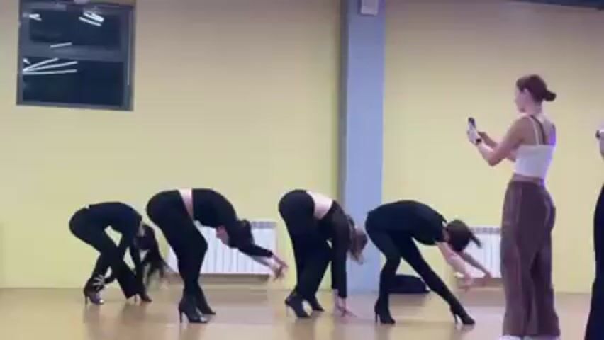 Four women dressed in black are practicing a dance routine. Another woman is filming them with her phone. The dance takes place in a studio with a window and a column. The walls are painted yellow.