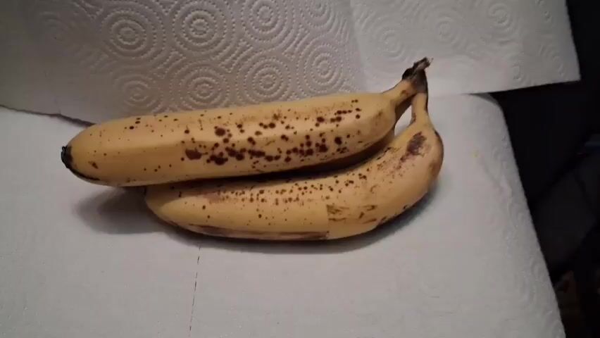The video displays two ripe bananas positioned side by side on a white paper towel. The bananas are yellow with brown spots, indicating ripeness. The background appears to be a surface covered with a textured white paper towel. The lighting is dim and appears to be artificial. There are no people or other objects visible in the frame. The camera angle is slightly from above, looking down at the bananas.