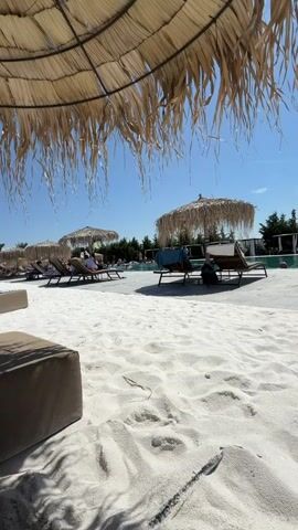The video pans across a sandy beach area with a swimming pool in the background. Several thatched umbrellas and lounge chairs are visible. The sand is white and appears soft. In the foreground, a dark brown ottoman or cushion is on the left. Towards the right, a flip-flop with orange and black accents rests on the sand. There are a few people lounging on chairs, some appearing to be reading or relaxing. The sky is clear and blue, indicating a sunny day. The overall scene depicts a resort or beach club atmosphere.