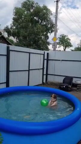 A child is playing in an inflatable pool with a green ball. The pool is blue and filled with water. The child is partially submerged in the water. The pool is surrounded by a white fence. There are trees and bushes in the background. A traffic sign is visible in the distance. The weather appears to be overcast.