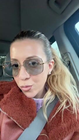 A woman in a car is filming herself. She is wearing sunglasses, a brown fuzzy coat over a lavender sweater, and a seatbelt. Her hair is blonde and pulled back in a ponytail. She smiles, then sticks her tongue out and makes a peace sign with her fingers. The car interior is visible. There is music playing in the background.