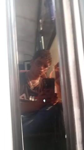 A person is taking a selfie in a mirror. The individual has curly hair and is wearing glasses, a black tank top with a logo, and a gold chain. The person appears to be puckering their lips. The mirror is framed with metal bars and shows a cluttered background. The person is holding a dark-colored smartphone, likely the camera being used for the selfie.