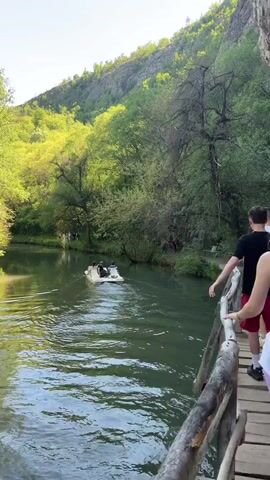 The video shows a group of people walking on a wooden bridge alongside a river. The river is surrounded by lush green trees and a mountain in the background. A small white boat with several people on board floats on the river. The wooden bridge has a simple wooden railing. The camera follows a woman wearing pink leggings, a white skirt, and a black backpack as she walks along the bridge. The bridge is built right up against a large rock face.
