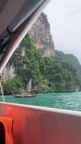 A video of a boat ride with a tropical island in the background. The island is covered in lush green trees and has steep cliffs. The water is a bright turquoise color. There is a longtail boat in the distance. The video is taken from the perspective of someone on another boat. The sky is cloudy.