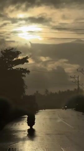 The video depicts a solitary figure riding a scooter on a wet, reflective road during what appears to be either sunrise or sunset. The sky is filled with dramatic, heavy clouds, illuminated by the bright sun peeking through. Tall trees and palm trees form a dense silhouette line in the background, suggesting a tropical or sub-tropical setting. Electrical wires and utility poles are visible along the side of the road, indicating a developed area.

The road surface is wet, likely due to rain, and the light reflects off it, creating a shimmering effect. The scooter rider is silhouetted against the bright sky, making their features indistinguishable. The overall mood of the scene is somewhat somber and contemplative, with the low light and wet conditions contributing to a feeling of isolation or peaceful journey. Other figures on motorcycles are visible further down the road, also silhouetted.

The time of day is either early morning or late evening, given the low angle of the sun and the cloud formations. The weather appears to be overcast with lingering moisture. The location is identified as Kathaluwa, Sri Lanka, which aligns with the presence of palm trees and tropical vegetation. There is no visible text in the video.