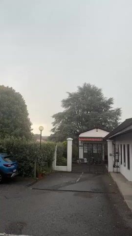 The media file displays a view of a driveway and entrance to a property on a cloudy day. A blue car is parked on the left side of the frame, facing away from the camera. To the right of the car is a dense green hedge, and a tall tree with a large canopy. In the background, a white gate with decorative elements is visible, leading to a building with a pink facade and a white trim. The sky is overcast, suggesting a cloudy or possibly rainy day. The overall mood is somewhat subdued, consistent with the gray sky. There are no people visible in the media file. The setting appears to be residential, possibly a private home or a small establishment, with elements like a lamppost and potted plants adding to the scene. There is no visible text in the media file.