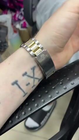 A close-up shot shows a person's wrist wearing a gold and silver digital watch with a metal band. The watch face is yellow and displays the time as 18:08:06. The person has a tattoo on their forearm. In the background, blurry objects are visible, including a striped item, a white bottle, and a black chair. The scene appears to be indoors.