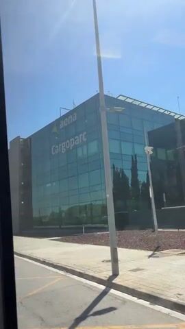 The media file shows a modern, multi-story building with a facade of reflective blue glass. The building is labeled with "aena Cargoparc." Aena is the Spanish airport operator, suggesting this is an airport cargo facility. The building appears to be situated in an industrial or commercial area, with a paved road and sidewalk in the foreground. Yellow lines mark the road. In the reflection of the glass, trees and a blue sky with a few wispy clouds are visible. The sunlight suggests it is daytime, likely a clear and sunny day. The perspective is from a moving vehicle, possibly a bus or car, as indicated by the framing and slight motion blur. There are no people visible. The overall impression is one of a functional, contemporary business environment.