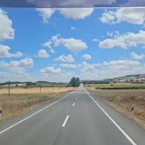 A straight, paved road with a white dashed line in the center and white lines on the edges cuts through a rural landscape. The road appears to be heading towards a slight incline in the distance. On either side of the road, fields of dry, yellow grass and crops stretch out. Clusters of green trees and vegetation are visible further back, as are rolling hills in the background. The sky is bright blue with scattered white, fluffy clouds. The video seems to be filmed from inside a moving vehicle, likely a car or bus, looking forward. There are no people or other vehicles clearly visible in the frame, but there are occasional small objects on the side of the road that might be signs or markers. The overall impression is a scenic drive through a warm, sunny day in the countryside.