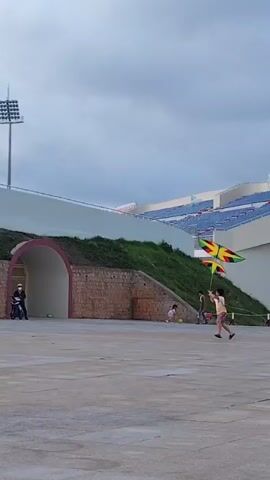 A young person is attempting to fly a colorful kite on a cloudy day. The kite, with green, yellow, and red colors, initially fails to launch and falls to the ground. The person is running across a gray paved area near a stadium with blue seating. A motorcycle is parked near an arched entrance in a stone wall behind them.  Another person is standing further away, watching the kite attempt. The scene appears somewhat deserted.