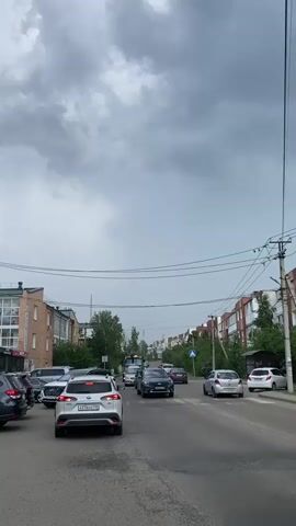 This is a video capturing a street scene in Markova, Russia. The primary subject is a street with several cars driving or stopped in traffic. The scene is set on a paved road lined with multi-story buildings, possibly apartment blocks, on both sides. Trees are visible between the buildings and along the roadside. Overhead power lines are prominent in the upper portion of the frame, crisscrossing the sky.

The time of day appears to be late afternoon or early evening, given the overcast and darkening sky. The weather is a significant element, with heavy, dark storm clouds dominating the sky, suggesting an impending storm or the aftermath of one. The overall mood is somewhat somber or dramatic due to the intense cloud cover.

The activity is the movement of vehicles on the road, with some cars appearing to be in a traffic jam. The camera is positioned at a height that suggests it's either from another vehicle or a person standing by the road.

Notable details include the license plates on some of the vehicles. The white SUV in the foreground has a license plate that appears to read "A376CA 138". Another car further down the road has a license plate that might be "C688HU 138". There's also a blue sign visible on the right side of the road, which is too blurry to read. A bus or large vehicle can be seen in the distance, moving through the traffic. There are no people clearly visible in the foreground, but the presence of vehicles implies human activity.