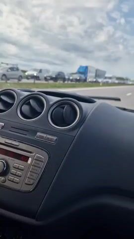A dashboard view from inside a vehicle shows a traffic jam on a highway. The sky is overcast with clouds. The dashboard is dark gray, with three circular air vents and a car stereo. An air freshener hangs from the rearview mirror. Outside, numerous vehicles including cars, vans, trucks, and trailers are lined up, barely moving.
