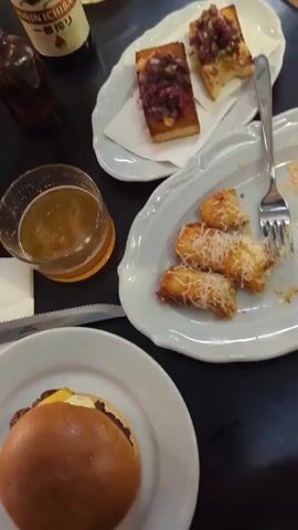 A video shows a dark-colored table top with several food and beverage items. A burger on a white plate is at the bottom left, along with a knife. A glass of beer and several plates with appetizers are arranged around the table. A bottle of beer and a small vase with flowers stand on the table. The camera moves and shows two bottles of beer on the table, as well as a couple of chairs in the background.