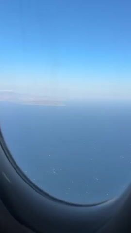 The video shows an aerial view from an airplane window. The scene is dominated by the vast blue expanse of the ocean, with a clear blue sky above. In the distance, a coastline with mountainous terrain is visible. Closer to the foreground, a densely populated coastal town with numerous buildings is seen. Several small, rocky islands are scattered in the water near the town. The edge of the airplane's wing and engine are partially visible along the right side of the frame. The ocean surface is choppy, with small whitecaps indicating waves. The sun appears to be shining brightly, casting a light over the scene.