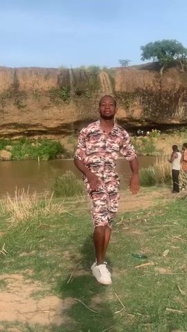 A man stands on a grassy area in front of a waterfall and a river. He is wearing a matching shirt and shorts with a camouflage pattern and white shoes. He is looking at the camera and smiling. There are other people in the background near the river. The sky is blue.