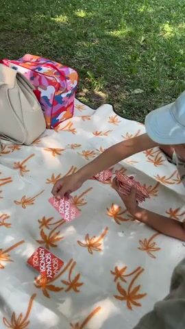 The video shows a person kneeling on a blanket spread on grass, playing a card game. The blanket has a pattern of orange palm trees. The person is wearing a light blue baseball cap and a blue shirt. They are holding a deck of cards in their left hand and placing a card on the blanket with their right hand. Several cards are already on the blanket, displaying the words "GONACK GONACK" in red and white text. To the left of the person, there is a cream-colored handbag and a colorful cooler bag with a pattern of pink, blue, and orange shapes. The background shows green grass and shadows cast by trees.
