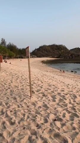 The video shows a sandy beach with a wooden pole and sign in the foreground. In the middle ground, a small group of people are sitting near the water. In the background, there is a hill covered in vegetation and the ocean. The sky is clear and light blue.
