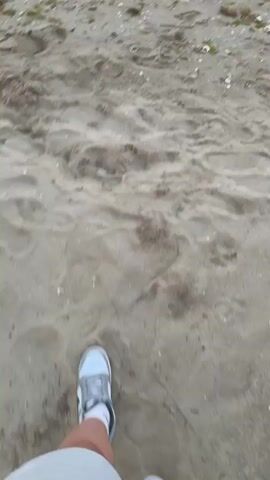The video shows a beach scene. The camera is focused on the sand and the water. There are footprints in the sand. The waves are gently lapping at the shore. The sky is a pale blue with hints of pink and purple. The overall impression is one of calm and tranquility.