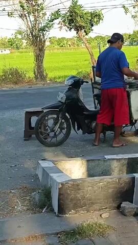 A man wearing a blue t-shirt and red shorts is standing next to a black motorcycle and a cart. He is looking down at the cart and appears to be preparing something. A motorcycle passes in the background from left to right at 00:01. Another person on a bicycle passes in the background from left to right at 00:04. The background shows a field of green crops and trees. The scene is outdoors, with a paved road and a concrete drainage area in the foreground.